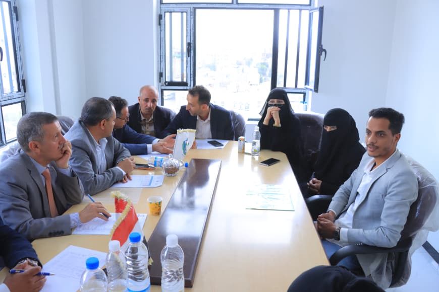 A mixed group of men and women in business attire sit around a long conference table in a bright room, discussing documents and beverages during a meeting.