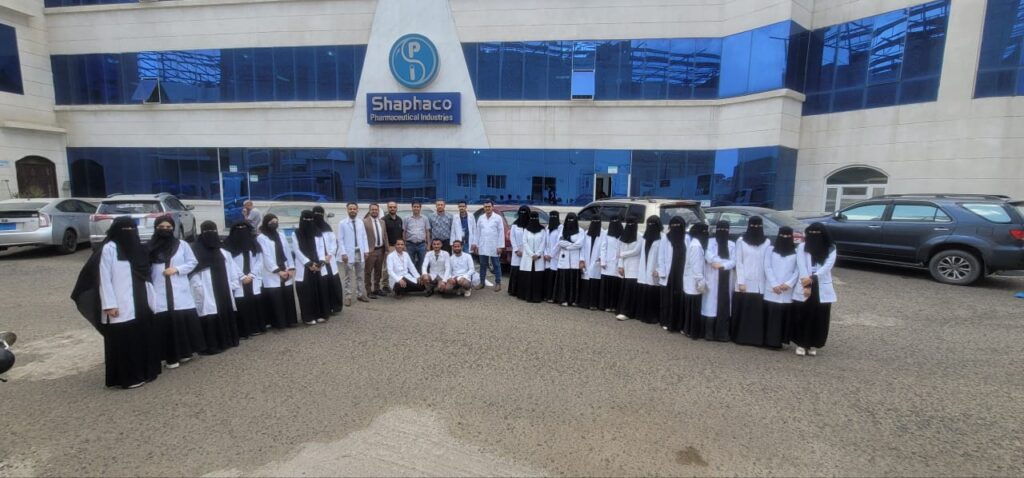Group of staff in white lab coats and black abayas posing outside the Shaphaco Pharmaceutical Industries building there are cars parked nearby