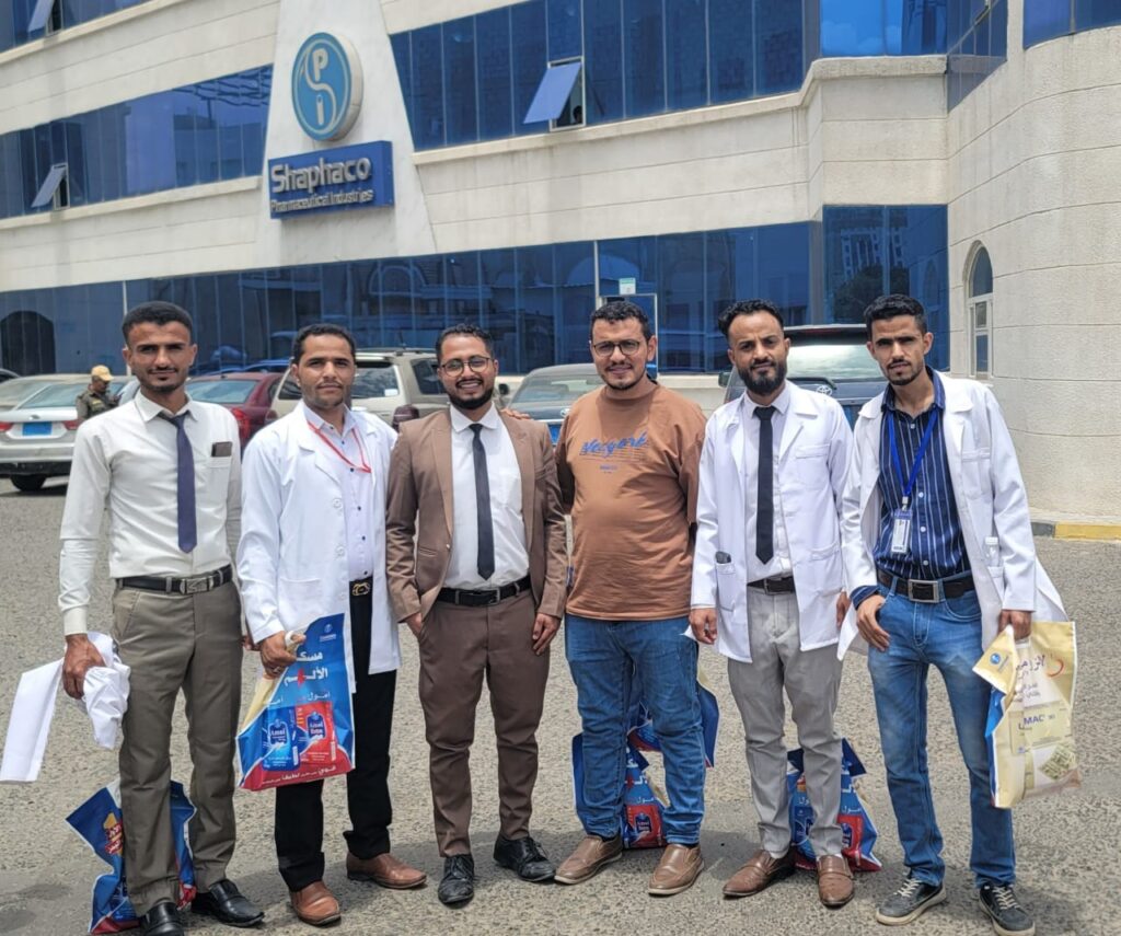 Six men stand outside a pharmaceutical building, some in white lab coats and others in business attire, holding bags.