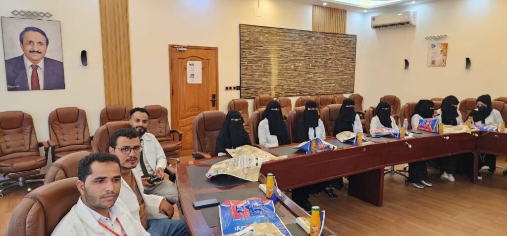 Group of people seated around a long conference table in a meeting room, some women wearing niqab, men in shirts and ties, with notes and drinks on the table.