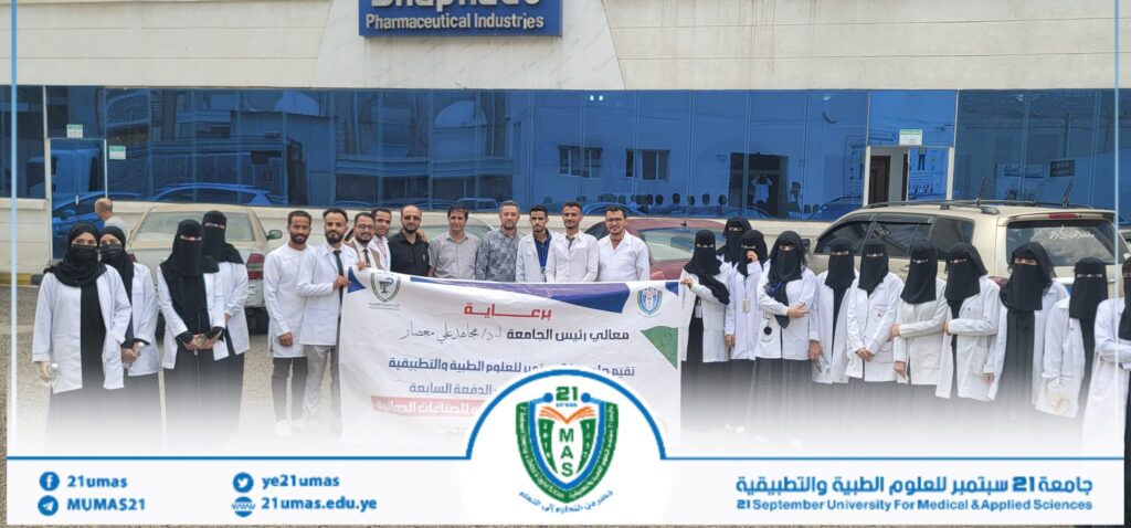 Group of students in white lab coats and hijabs posing with a large banner in front of a university building with blue-tinted windows.