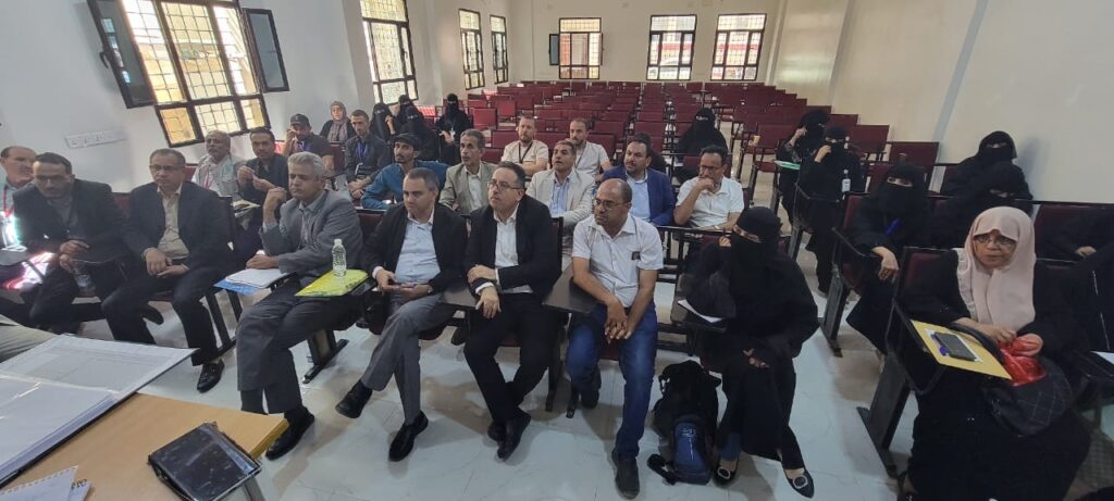 Audience seated in a lecture hall for a meeting or seminar; front row mostly men in suits, several women in black hijabs/niqabs, notebooks and folders visible.