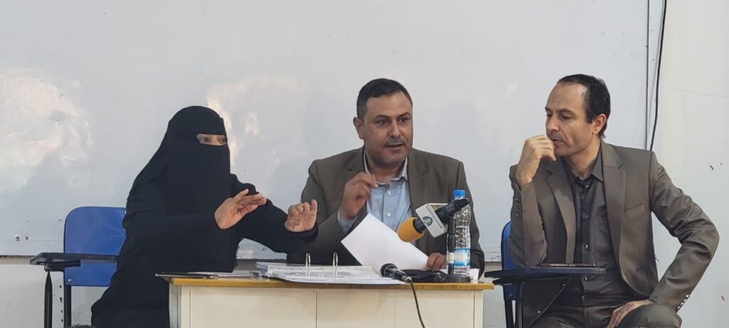 Woman in a black niqab speaks at a panel desk with two men; microphones, papers, and a water bottle on the table.