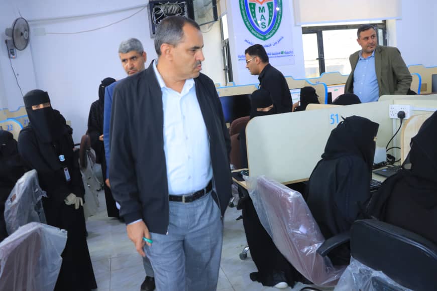 Man in a light blue shirt and dark jacket walks through a busy call-center-like office with women in black abayas at computer desks behind him.