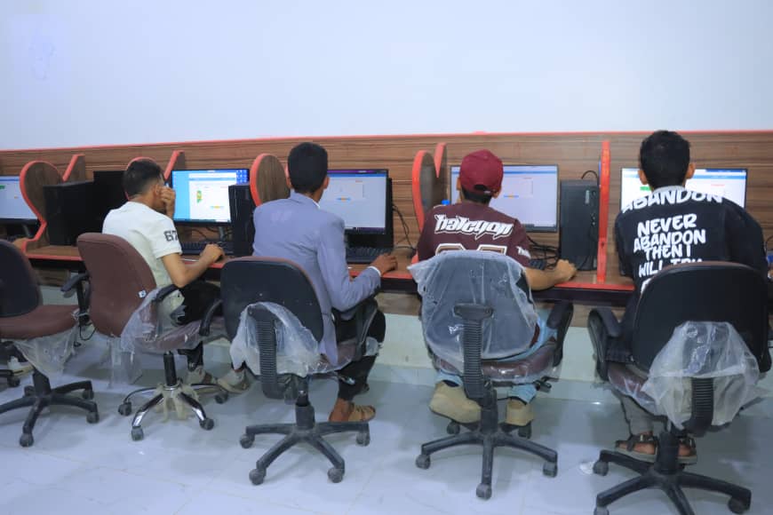 Group of students sitting at a row of desktop computers in a classroom, focused on screens, backs to the camera.