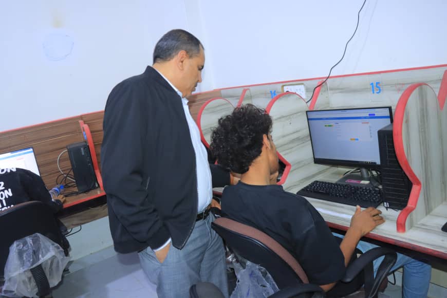 Supervisor stands beside a student at a desktop computer in a row of computer stations in a lab setting.