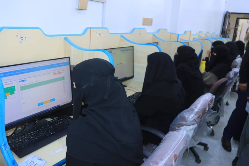 Women in black abayas seated at computers in a row of study cubicles, focusing on screens in a computer lab.