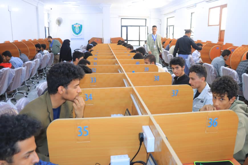 Crowded computer lab or exam hall with students at wooden numbered cubicles (34–38); a supervisor walks in the back of the room.