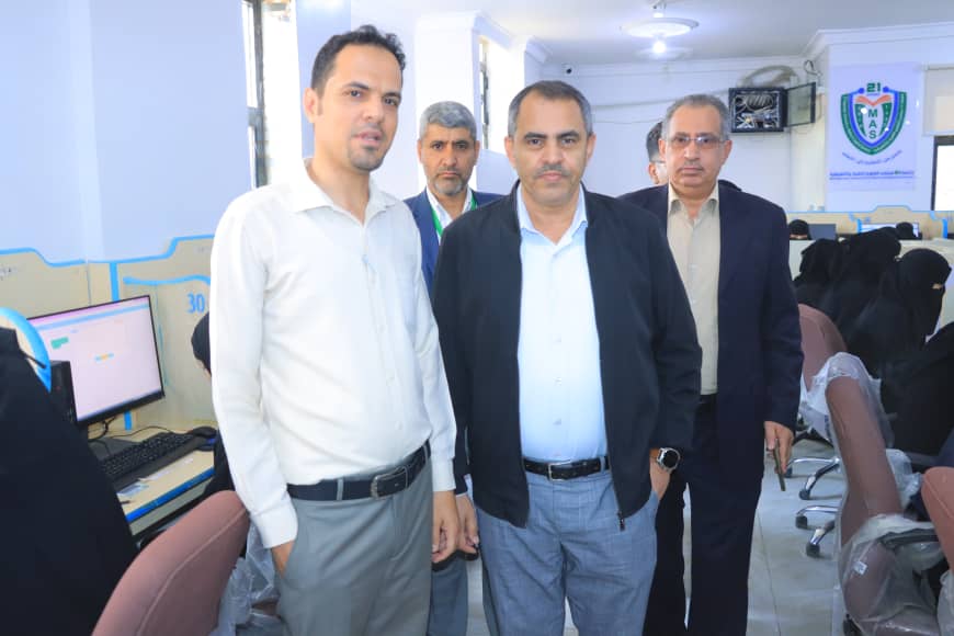 Four men stand together in a computer lab or training room with desks and monitors visible in the background.