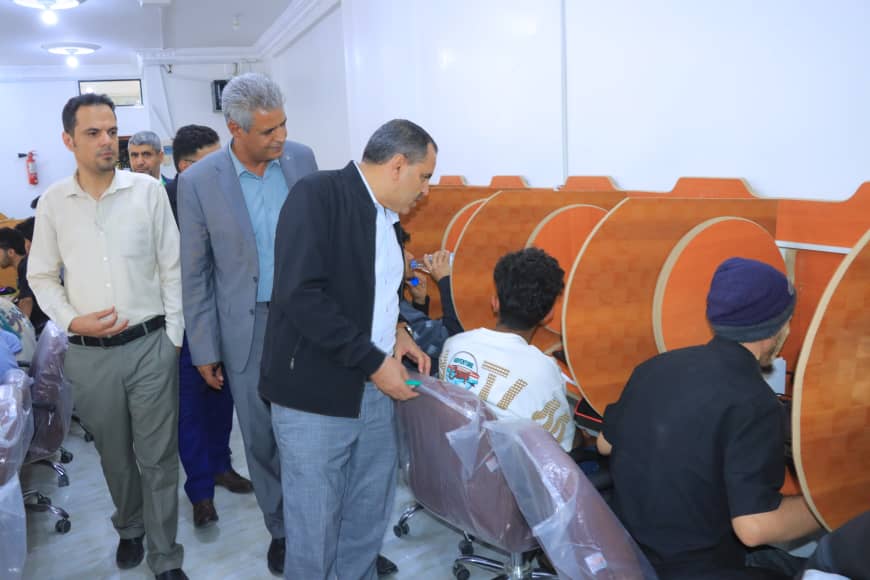 Group of men in business attire walking through a computer lab while students sit at orange curved desks.