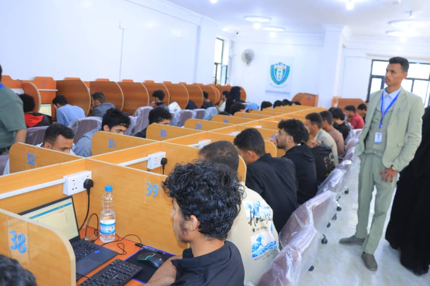 Rows of wooden cubicles with students using laptops in a computer lab; a supervisor in a light suit stands nearby.