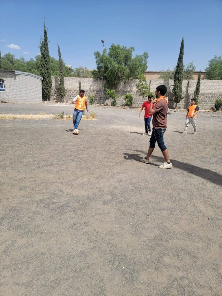 Group of young men playing soccer on a dusty outdoor lot; one player in orange controls the ball while others watch on a sunny day.