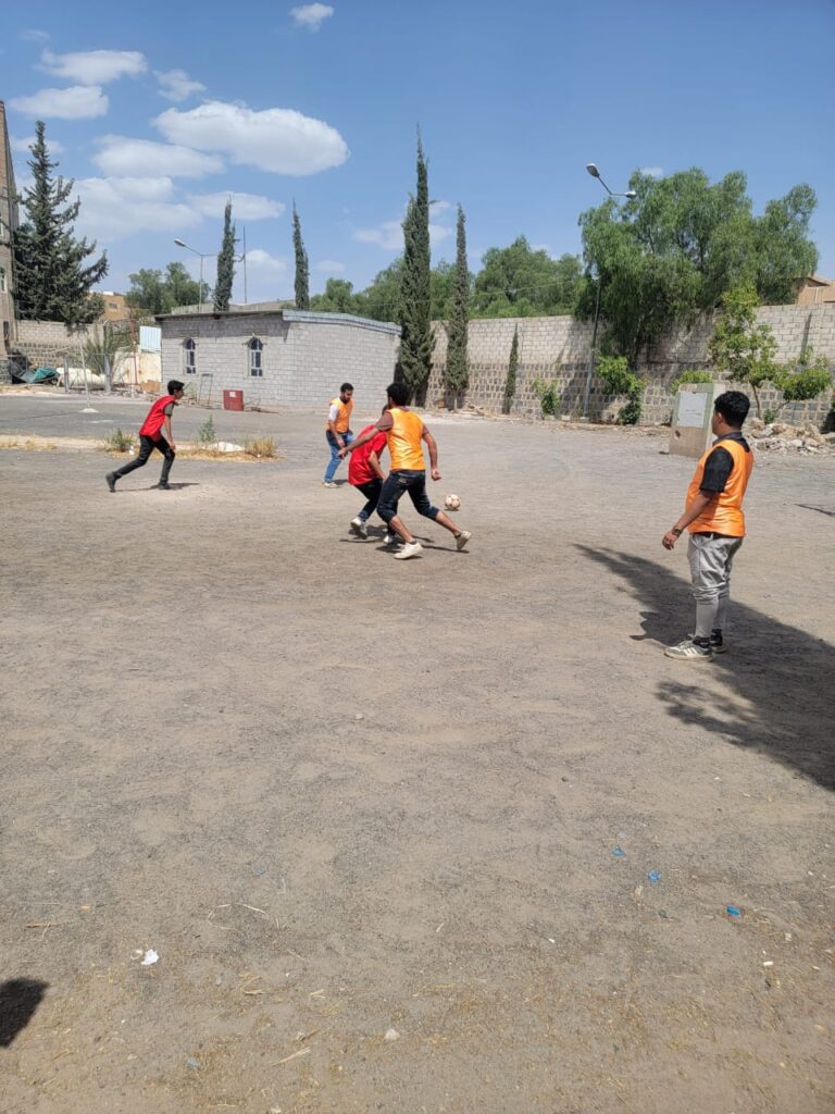 Group of young players on an outdoor dirt field, playing soccer with an orange pinnie vest nearby a wall and trees.