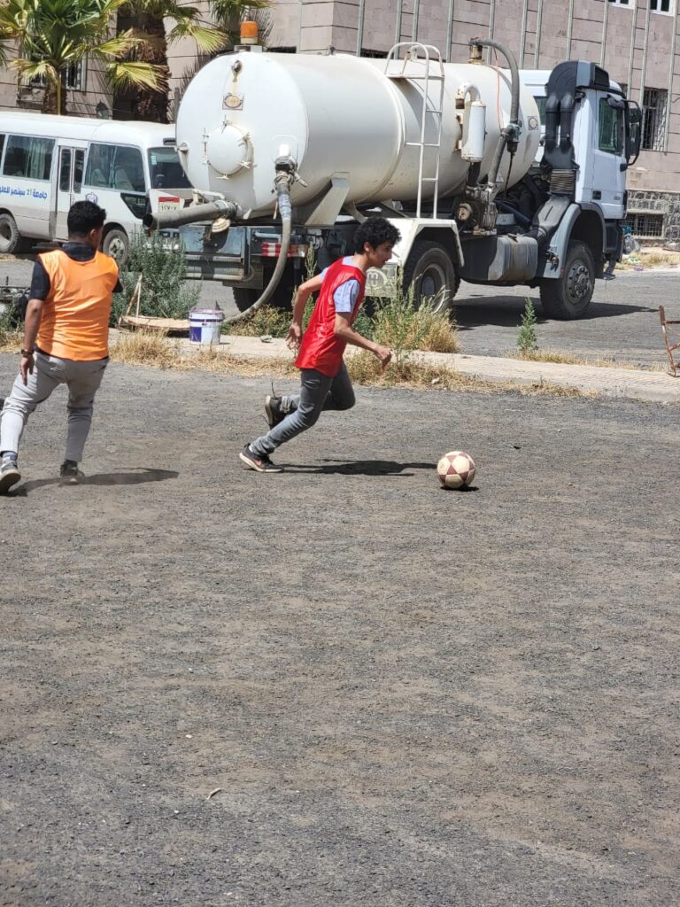 Two boys in orange and red vests play soccer on a dusty lot, with a large white tanker truck in the background.