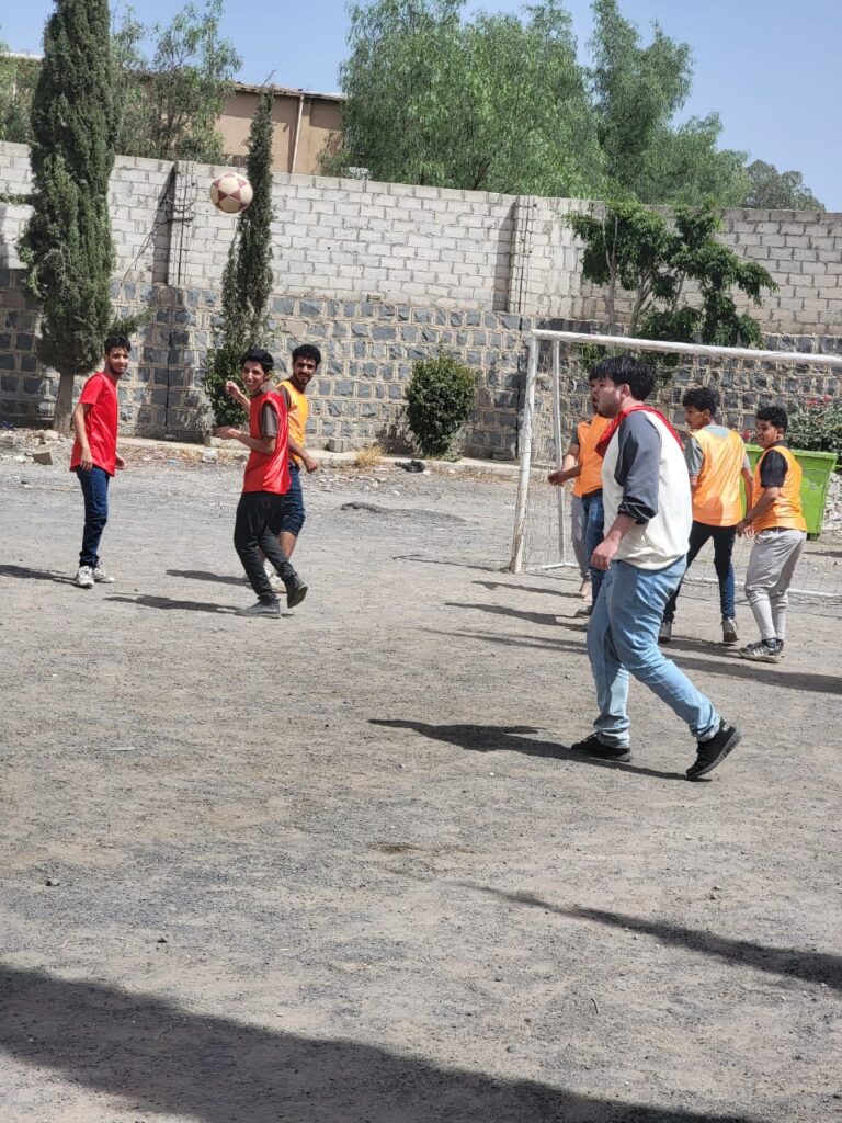 Group of young men playing soccer on a dusty outdoor field, ball in midair near a goalpost against a stone wall.