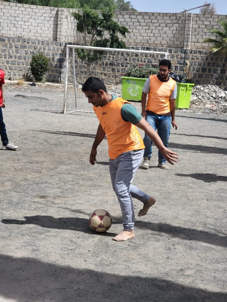 Man in an orange jersey kicks a soccer ball on a dusty field as a teammate watches; a goal and green dumpsters are in the background.