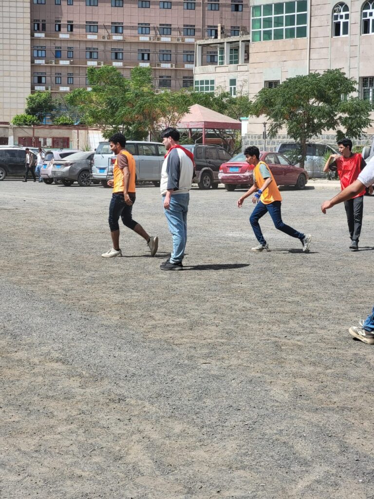 Group of young men running and playing on a gravel parking lot outside a campus building, some wearing orange vests.
