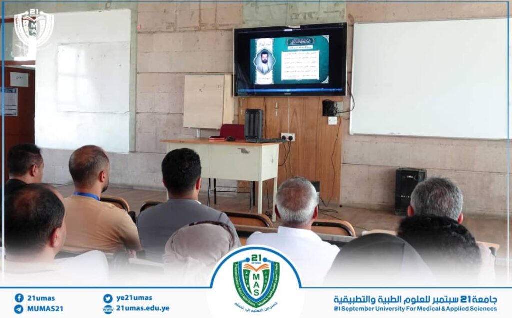 1001171676 - جامعة 21 سبتمبر Adult students seated in a classroom watching a presentation on a wall-mounted screen; desk with projector equipment in front.
