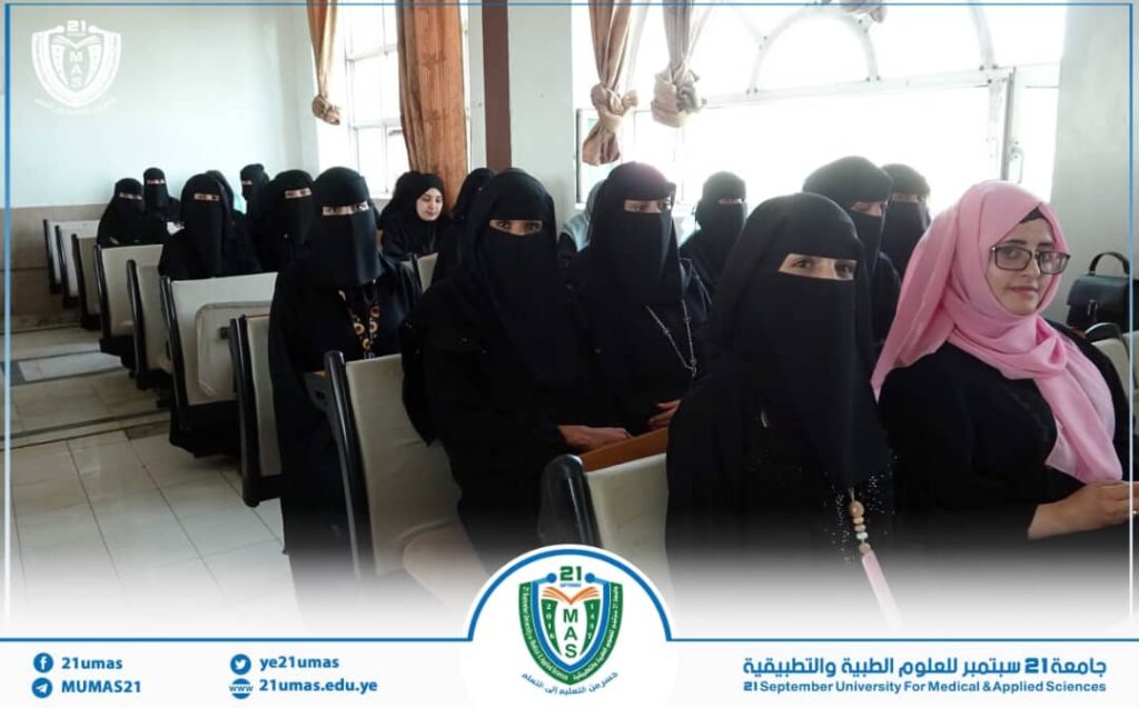 1001171683 - جامعة 21 سبتمبر Group of women in black hijabs seated in a classroom; a woman in a pink hijab sits at the front, with university logos visible.
