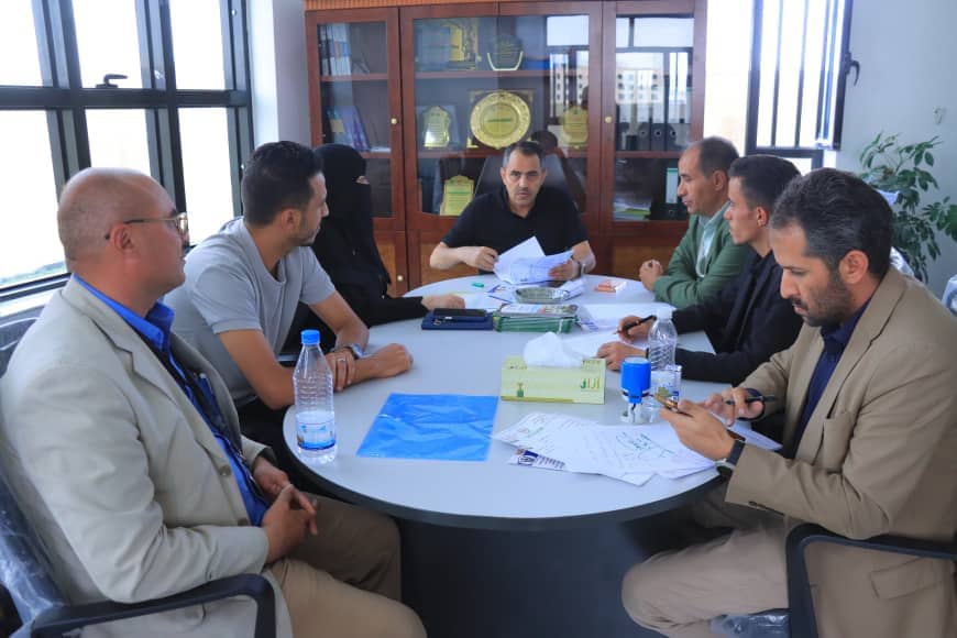 Seven professionals around a round conference table reviewing papers in a meeting room.