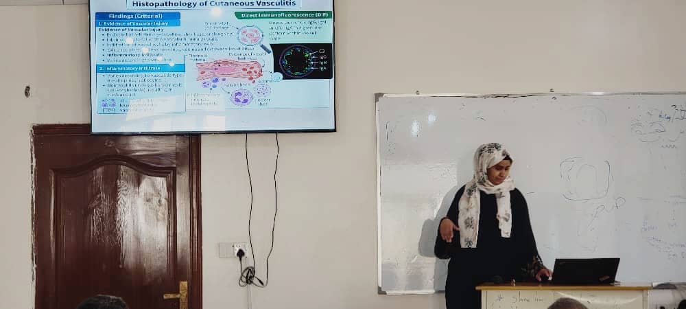 Female instructor wearing a hijab gives a lecture beside a whiteboard and a laptop in front of students; a slide about histopathology is displayed on a screen above the door.