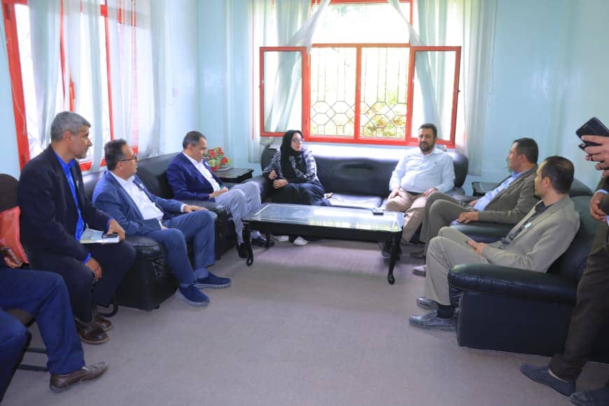 Eight adults seated on black leather couches around a glass coffee table in a bright, blue-walled living room with red window frames. A woman wearing a hijab sits among seven men in a semicircle, engaged in discussion. Group discussion in a sunny room with light curtains and a central glass table. Professional meeting taking place in a home setting, with attendees facing each other on couches. Informal conference-style gathering of colleagues in a cozy living-room environment.