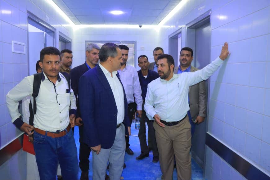 Group of men in business casual in a white-tiled hallway, with one man gesturing toward the wall as others listen.
