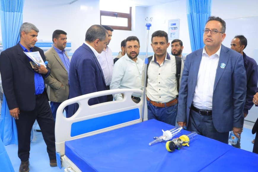 Group of men in business attire stand around a hospital bed with blue sheets in a clinical room.