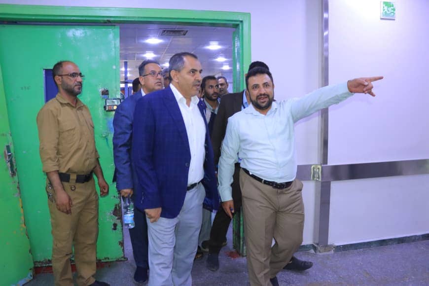 Group of men walking through a doorway; man in light blue shirt points to the right while others follow and look in that direction in a building corridor.