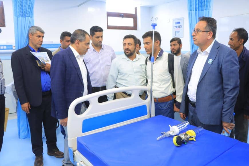 Group of men in business attire gather around a hospital bed in a bright room, with an IV pole and medical equipment on a blue-covered table nearby.