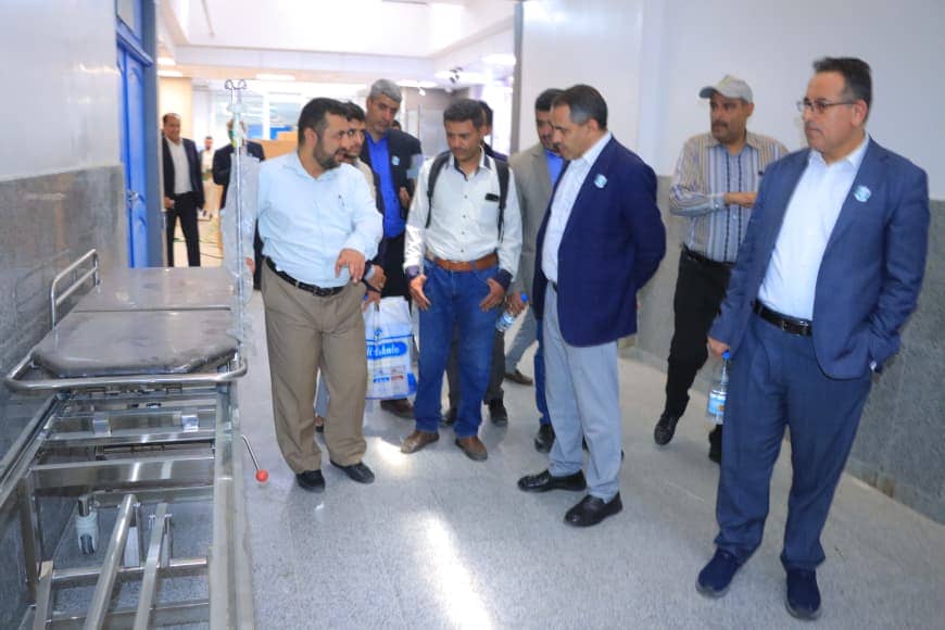 Group of men in business attire touring a hospital corridor with medical equipment carts and IV stand.