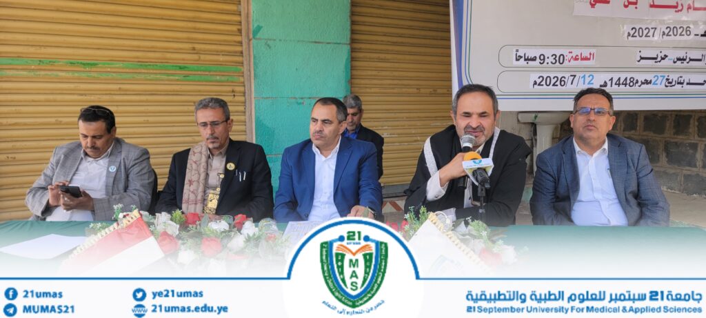 Panel of men in suits seated at a table during a university event, one man speaking into a microphone; Arabic banner behind them and the university emblem visible at the front.
