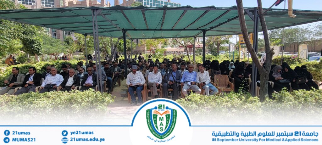 Audience seated under a green canopy at an outdoor university event, with many attendees in formal attire and women in black abayas; university logo banner at bottom.