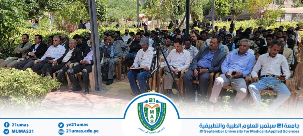Large outdoor conference scene with dozens of attendees seated in rows under a pavilion, some taking notes, a camera on a tripod near the center, and a university banner at the front.