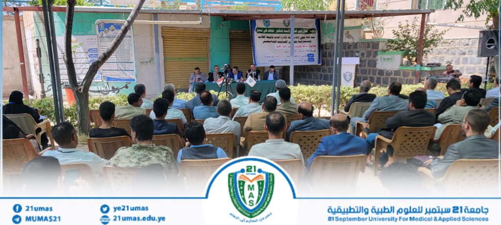 Outdoor panel discussion with six speakers at a long table under a canopy, addressing a seated audience with banners behind them and a university emblem visible on the stage backdropUI.
