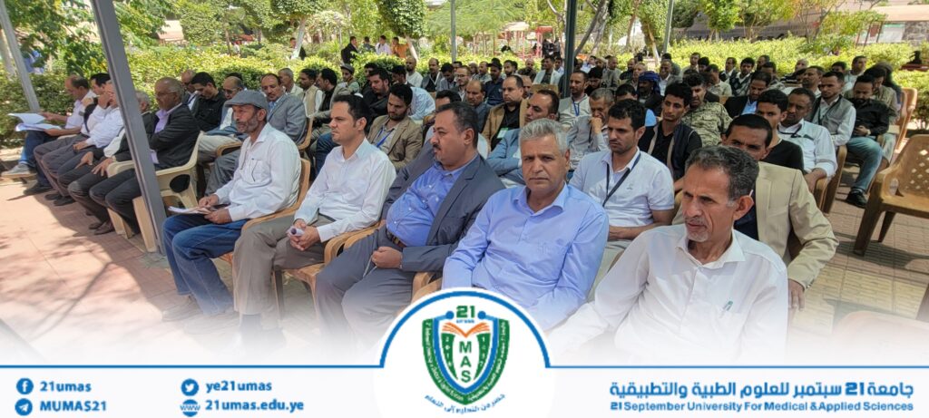 Audience of men seated in rows at an outdoor university seminar, with greenery and shade structures in the background and a campus banner at the bottom.