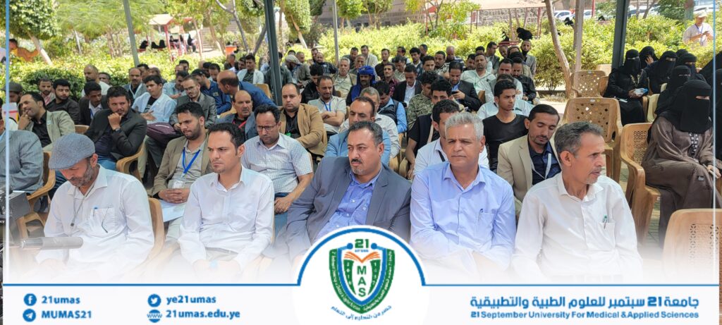 Crowd of people seated outdoors at a university event, with a banner and seal visible at the bottom and trees in the background.