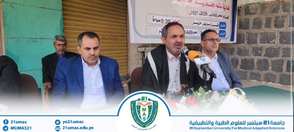 Panel of men seated at an outdoor press conference, central speaker with a microphone, others beside him; banner with Arabic text behind and a university logo at the bottom.