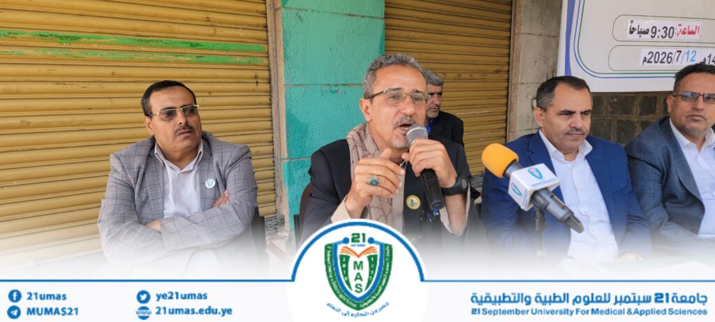Panel of men at a press conference, central speaker holds a microphone with a university emblem nearby.