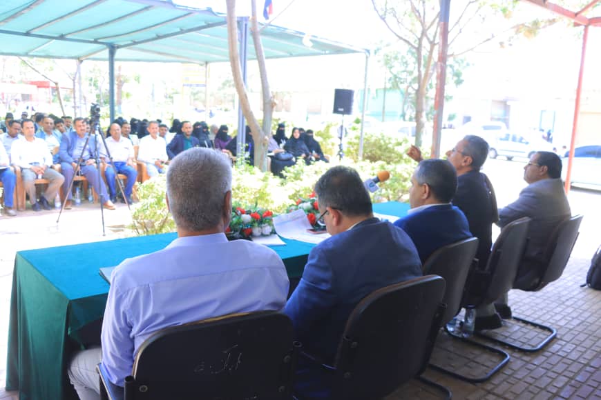 Panel of five men in suits seated at a green table under a canopy addressing an outdoor audience.