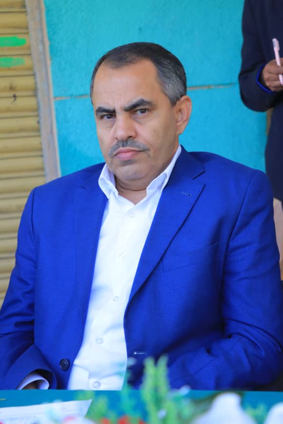 Middle-aged man in a blue suit and white shirt seated at a table, looking toward the camera with a serious expression, teal wall background.