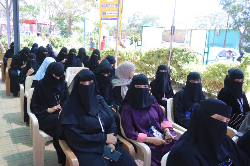 Group of women wearing niqabs seated in plastic chairs at an outdoor event.