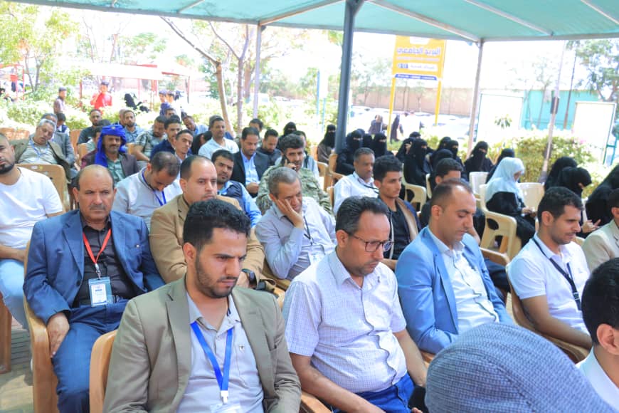 Audience seated in rows at an outdoor conference under a green shade canopy, many wearing lanyards and badges.