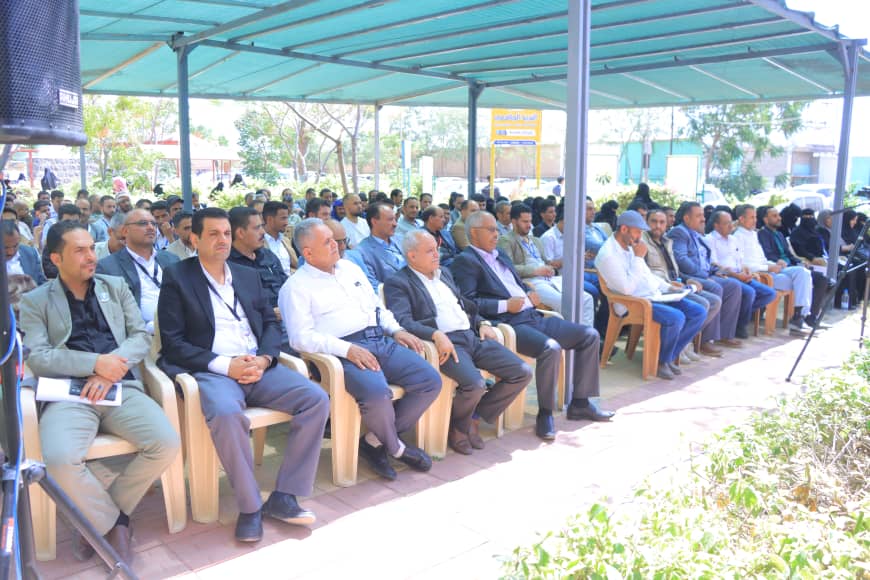 Group of professionally dressed people seated in rows under a blue canopy at an outdoor conference, listening to a speaker on a sunny day.