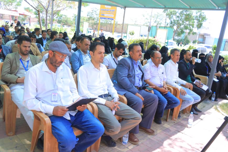 Audience seated outdoors under a green canopy at a conference, many wearing lanyards and business attire.