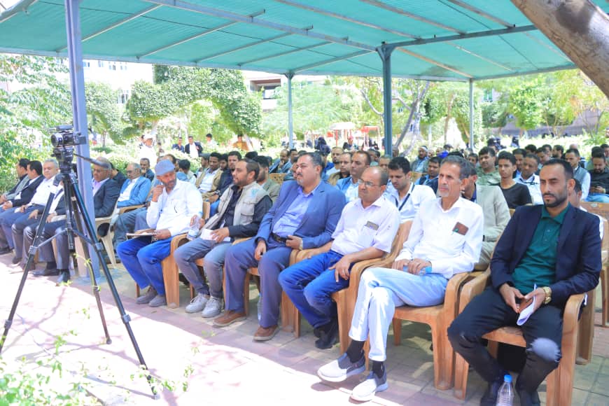 Group of men seated under a turquoise canopy at an outdoor event, with a camera on a tripod nearby.