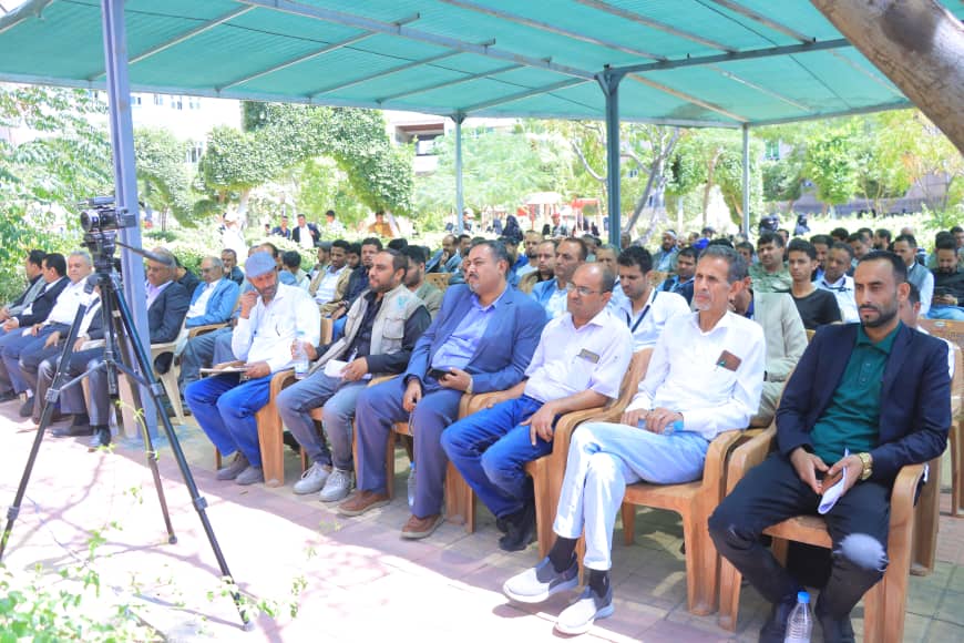 Audience seated under a teal canopy at an outdoor event, with a video camera on a tripod filming nearby.