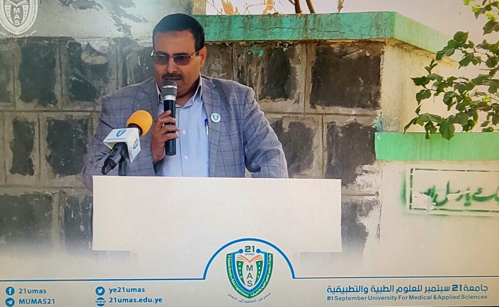 Man in a blazer and glasses speaks into a handheld microphone at a podium with a university crest behind him in an outdoor setting.