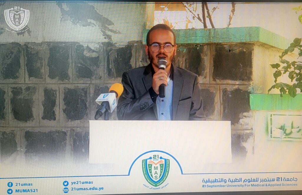 Man in a suit speaks into a microphone at a lectern, with a university crest on the podium behind him.