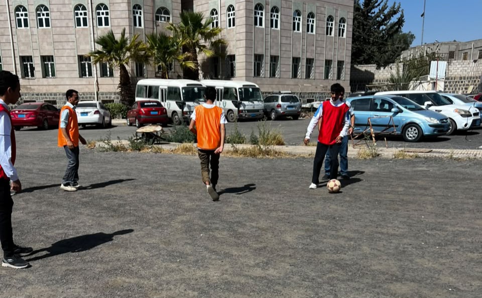 Group of young men playing casual soccer in a parking lot near a campus building; two players wear orange vests, one with the ball nearby.
